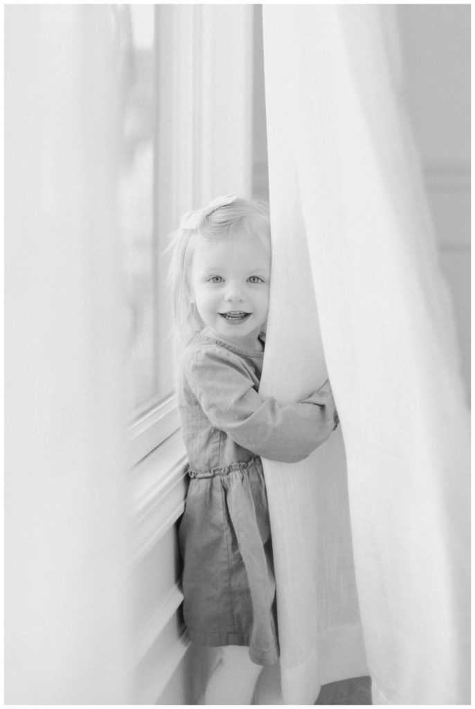 A black and white photo of a toddler girl standing by the window smiling at the camera while hiding behind the white curtains. 