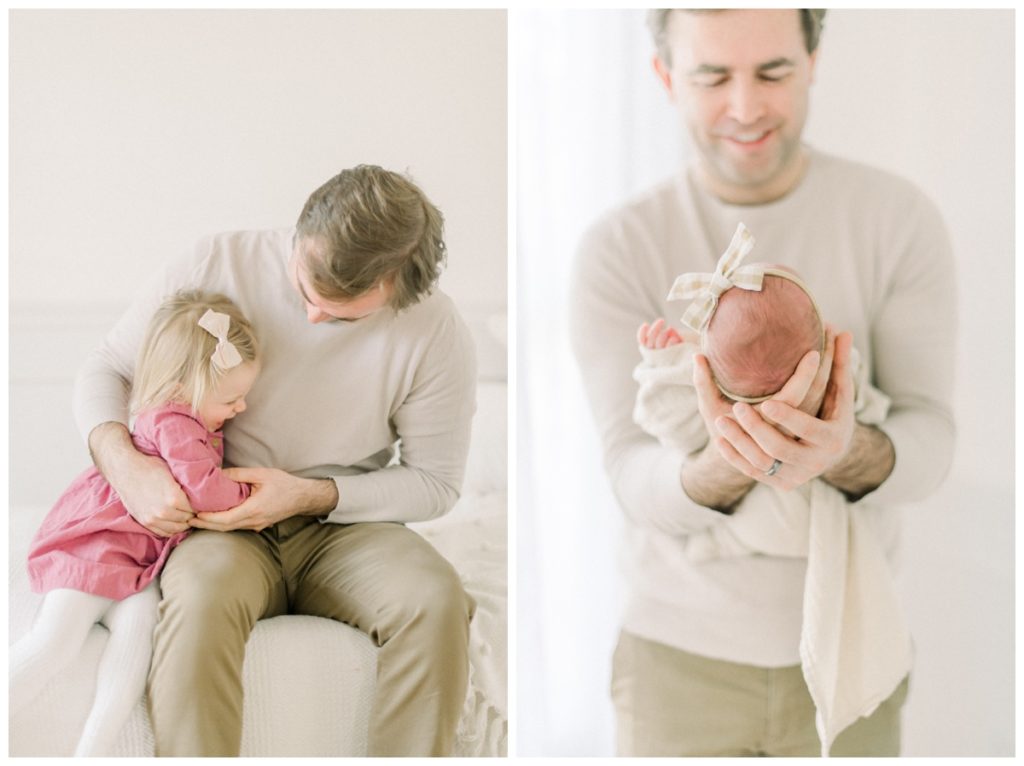 Photo of a dad standing in a white natural lit studio holding his newborn daughter to his stomach with both hands.